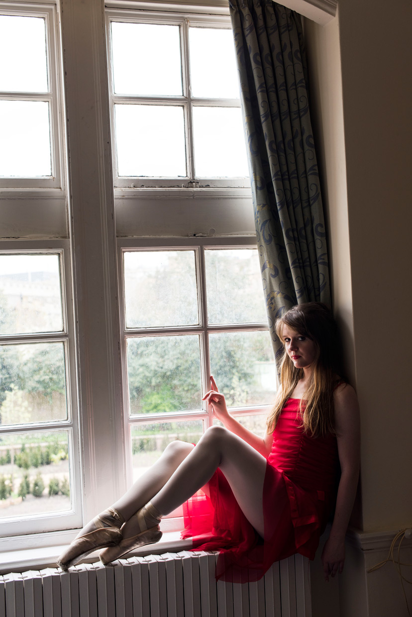 Naomi Grace in red dress and ballet shoes sitting on windowsill at Christ's College, Cambridge.
